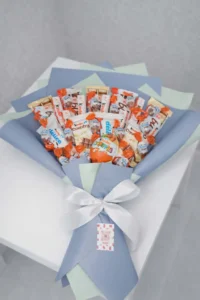 Handcrafted sweet bouquet featuring Kinder Surprise eggs, Kinder Bueno, and Delice bars wrapped in pastel blue and mint paper with a white satin ribbon.