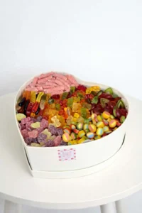 White heart-shaped gift box filled with a colorful assortment of jelly candies, sour sticks, and gummy bears