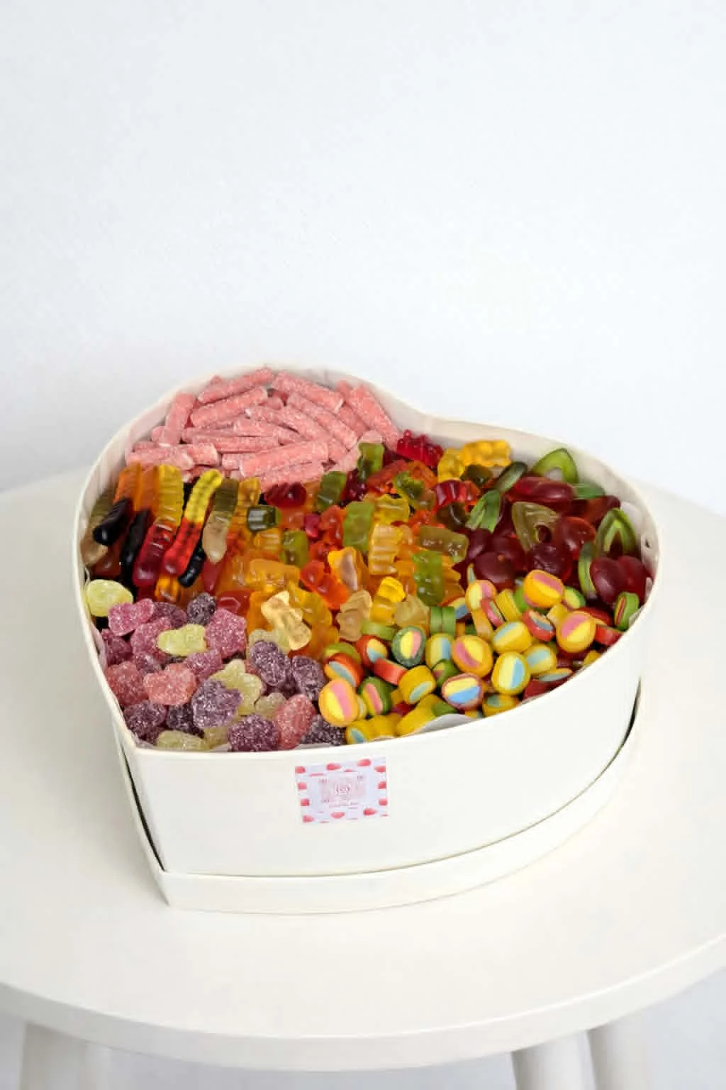 White heart-shaped gift box filled with a colorful assortment of jelly candies, sour sticks, and gummy bears