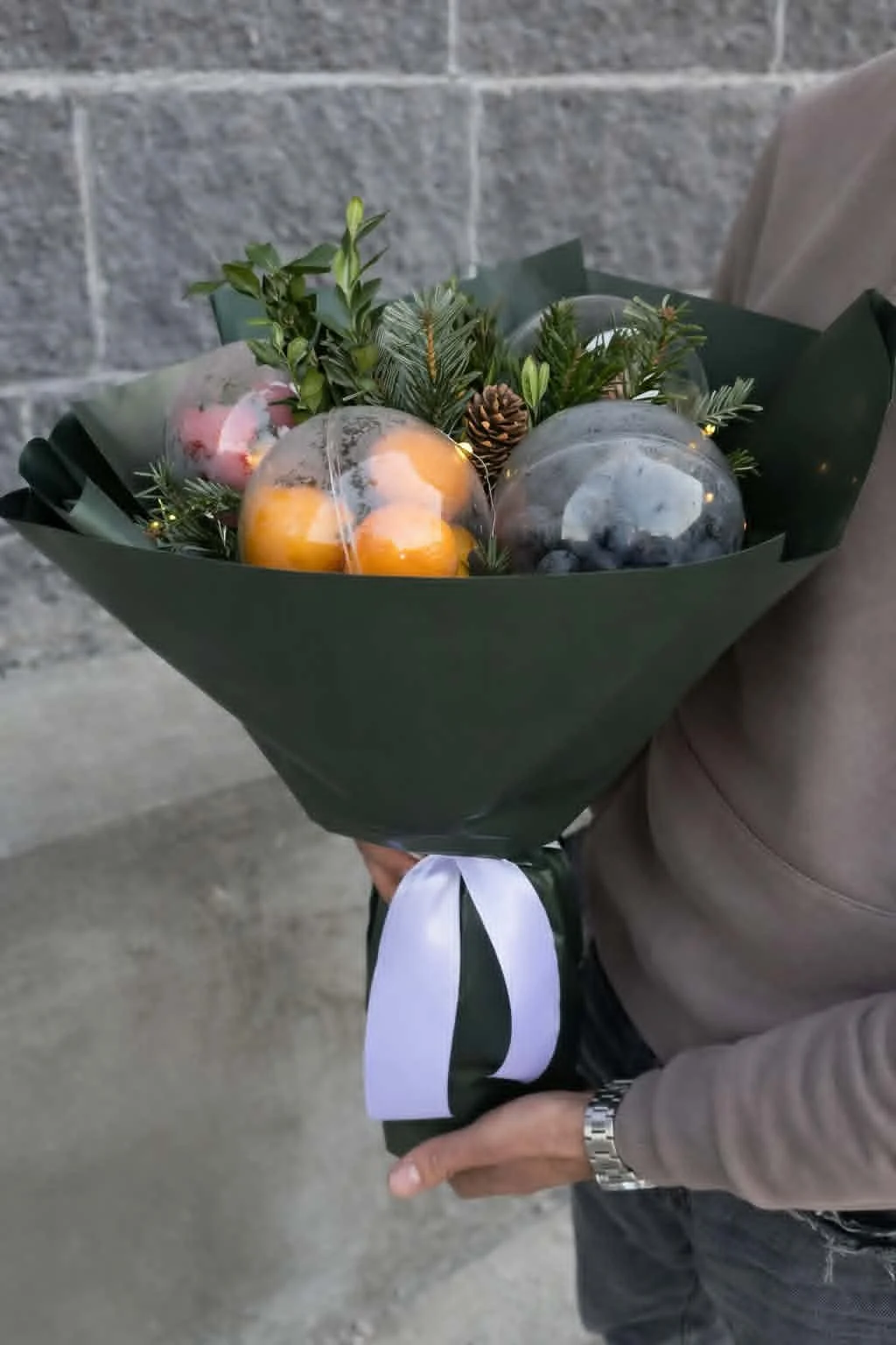 Premium winter edible gifts arrangement in a handheld bouquet of tangerines, nuts, and evergreen boughs in dark green paper wrapping.