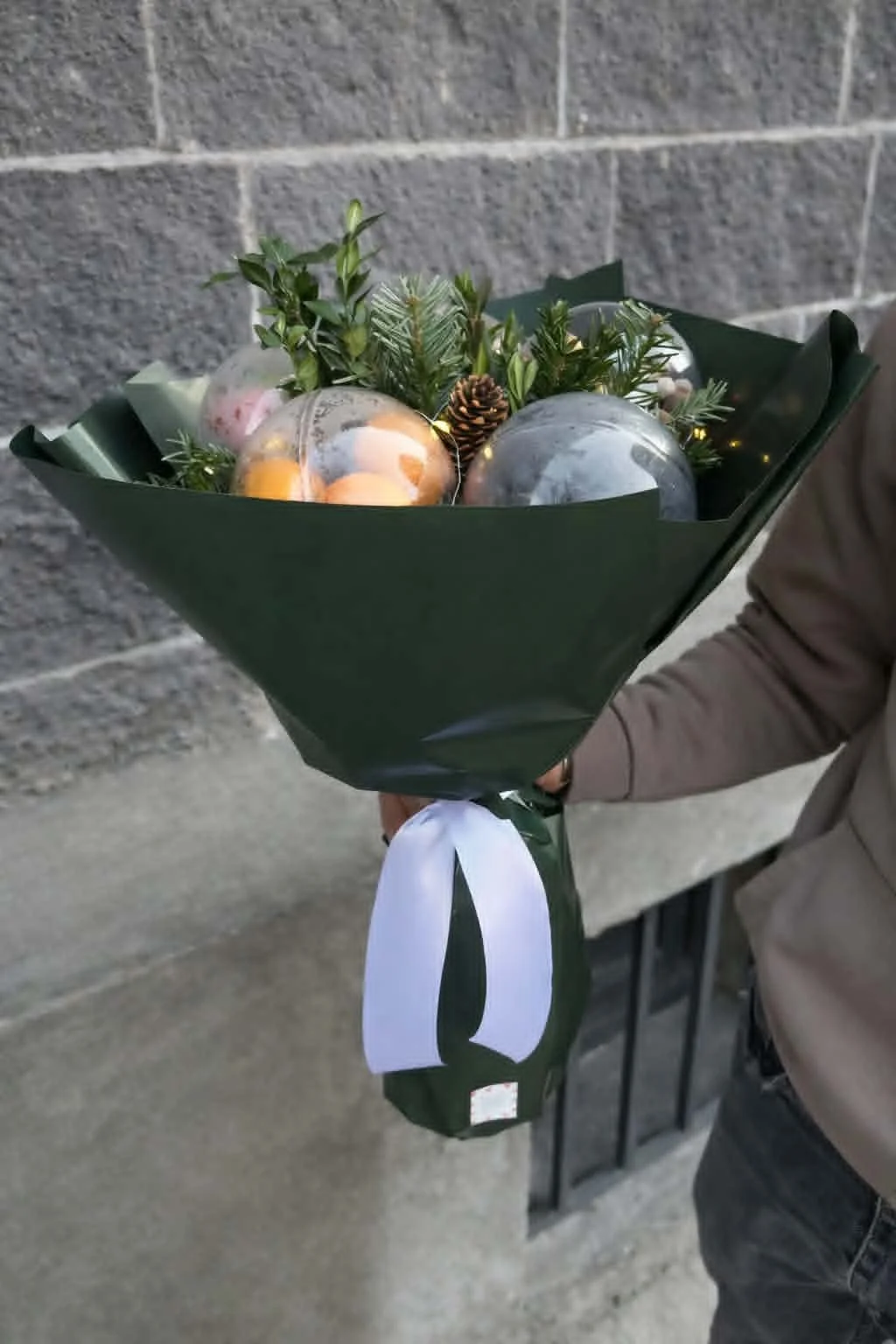 Premium winter edible gifts arrangement in a handheld bouquet of tangerines, nuts, and evergreen boughs in dark green paper wrapping.
