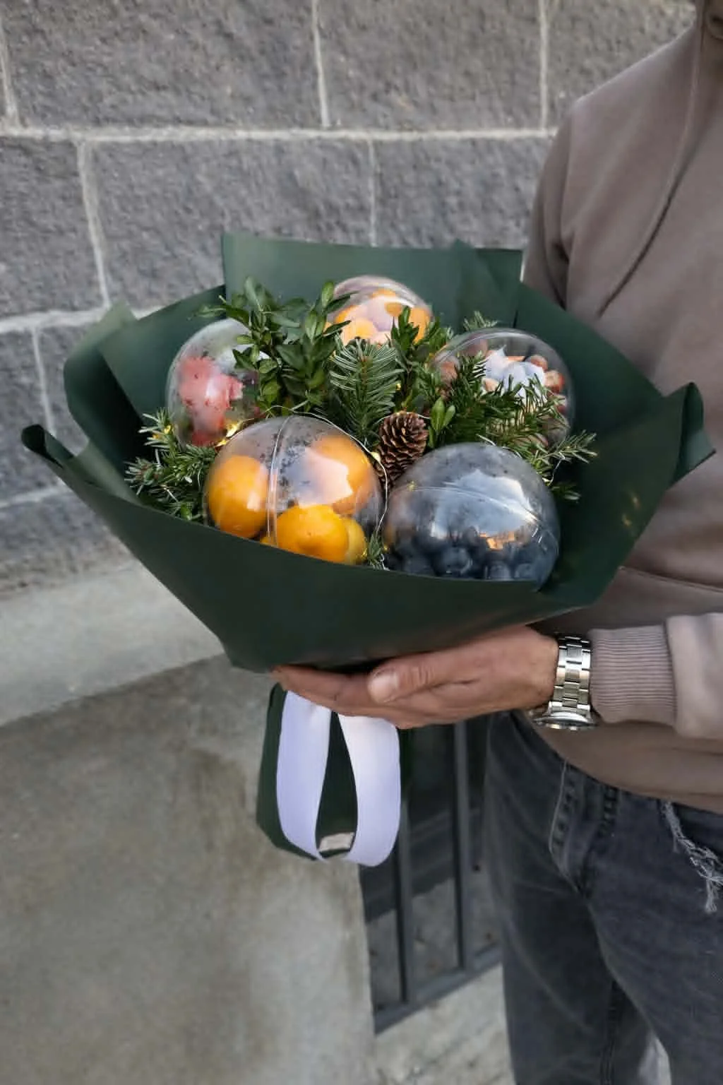Premium winter edible gifts arrangement in a handheld bouquet of tangerines, nuts, and evergreen boughs in dark green paper wrapping.