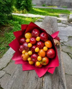 BooqArt's Signature Fresh Fruit Gift Bouquet resting on a rustic weathered wooden bench, featuring glossy red apples and vibrant orange stone fruit in pointy red paper wrapping with a brown ribbon.
