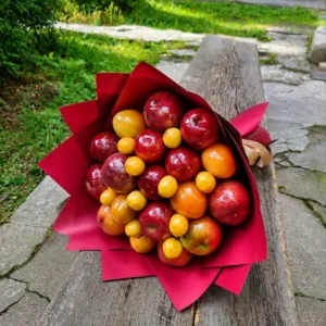 BooqArt's Signature Fresh Fruit Gift Bouquet resting on a rustic weathered wooden bench, featuring glossy red apples and vibrant orange stone fruit in pointy red paper wrapping with a brown ribbon.