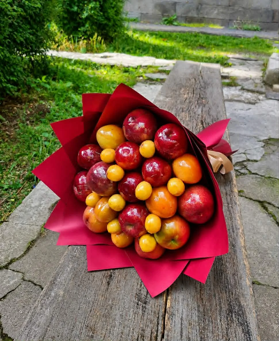 BooqArt's Signature Fresh Fruit Gift Bouquet resting on a rustic weathered wooden bench, featuring glossy red apples and vibrant orange stone fruit in pointy red paper wrapping with a brown ribbon.