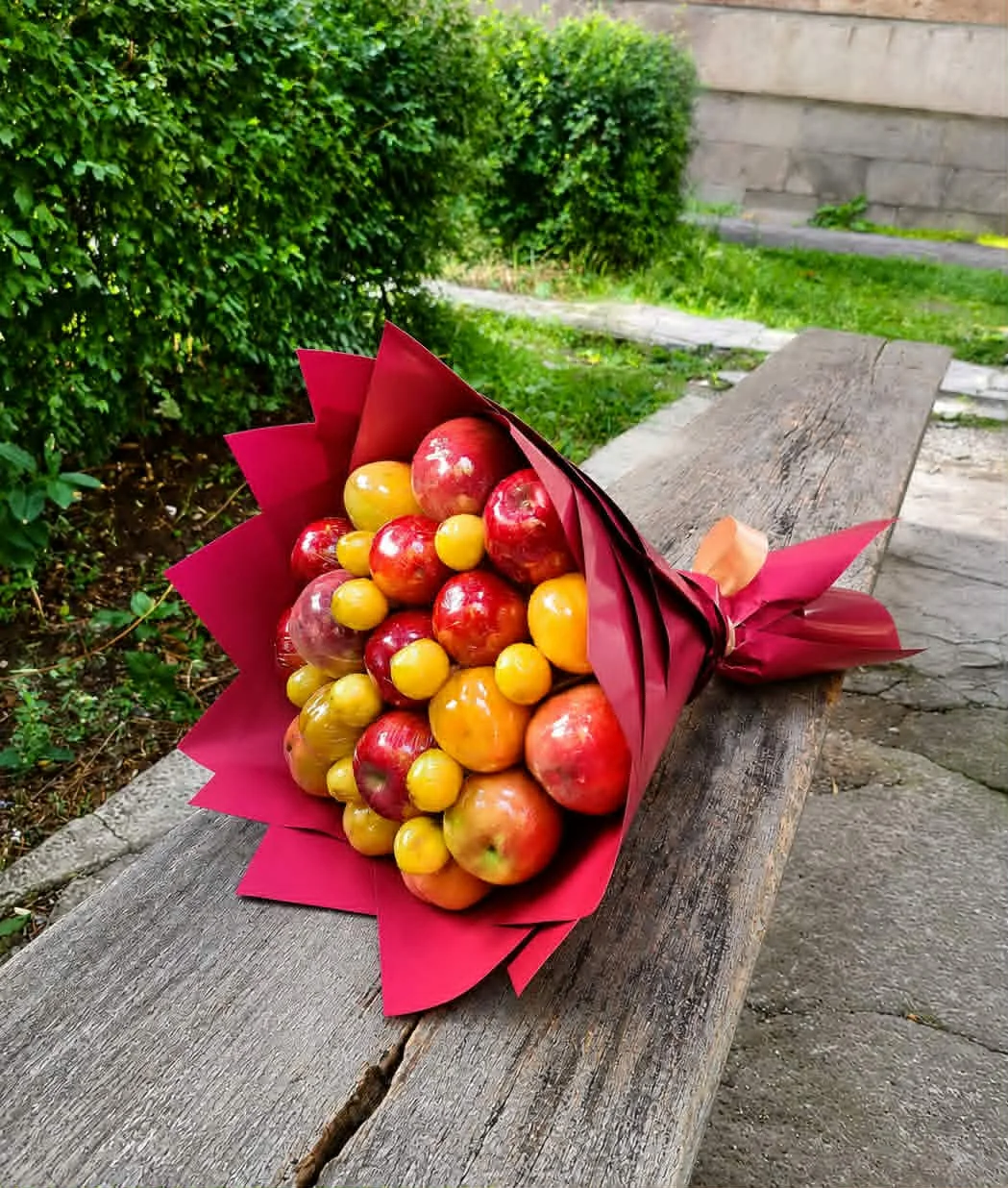 BooqArt's Signature Fresh Fruit Gift Bouquet resting on a rustic weathered wooden bench, featuring glossy red apples and vibrant orange stone fruit in pointy red paper wrapping with a brown ribbon.